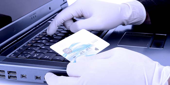 Man wearing rubber gloves, holding a credit card and typing the card numbers into a laptop